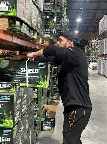 A man in a black hoodie and black cap measures a box of Aloe SHIELD hand distill gloves in a warehouse aisle, with shelves stacked with product boxes on both sides.