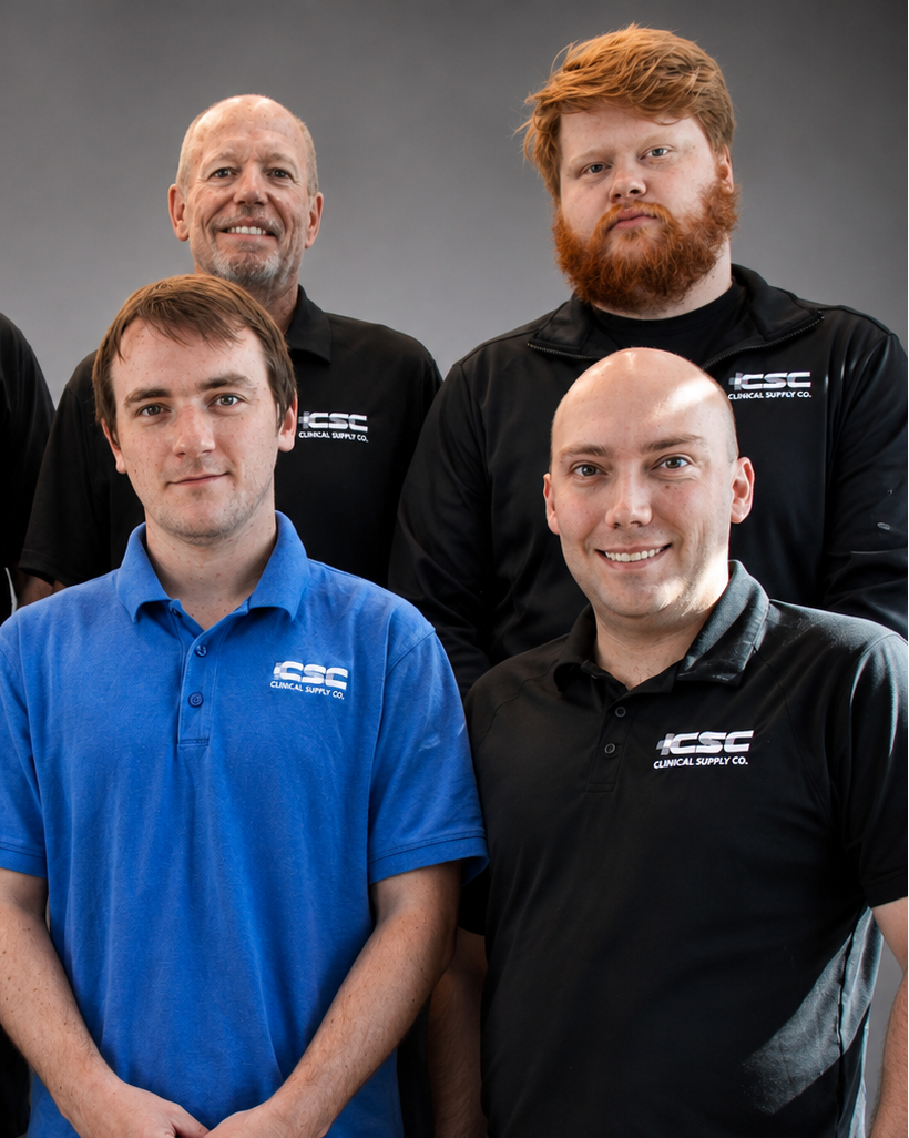 A group of six men posing together in a studio setting, wearing matching black polo shirts with a white 'CSC' logo on the chest. One man in the center wears a bright blue polo shirt.